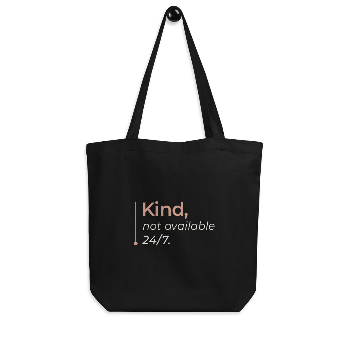 A tote bag preta Tag&Get com a frase “Kind, not available 24/7.” em tons claros e pêssego, pendurada pelas alças num gancho sobre fundo branco.