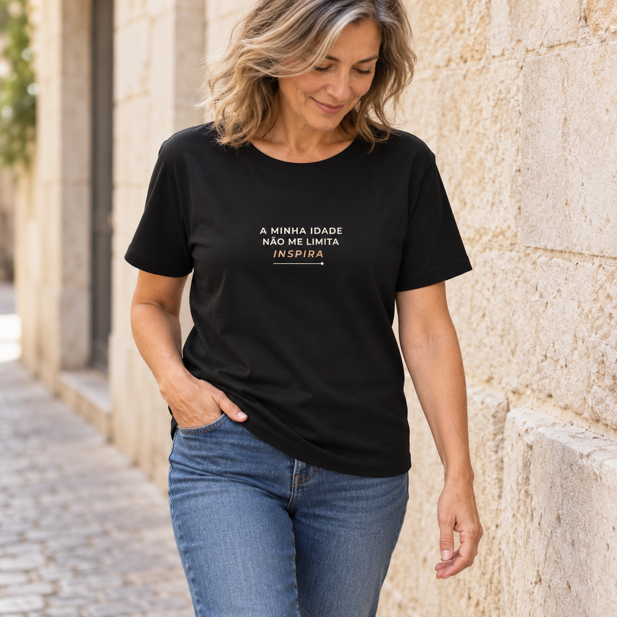 Mulher com T-shirt preta «A minha idade não me limita, inspira» e jeans, junto a uma parede de pedra.