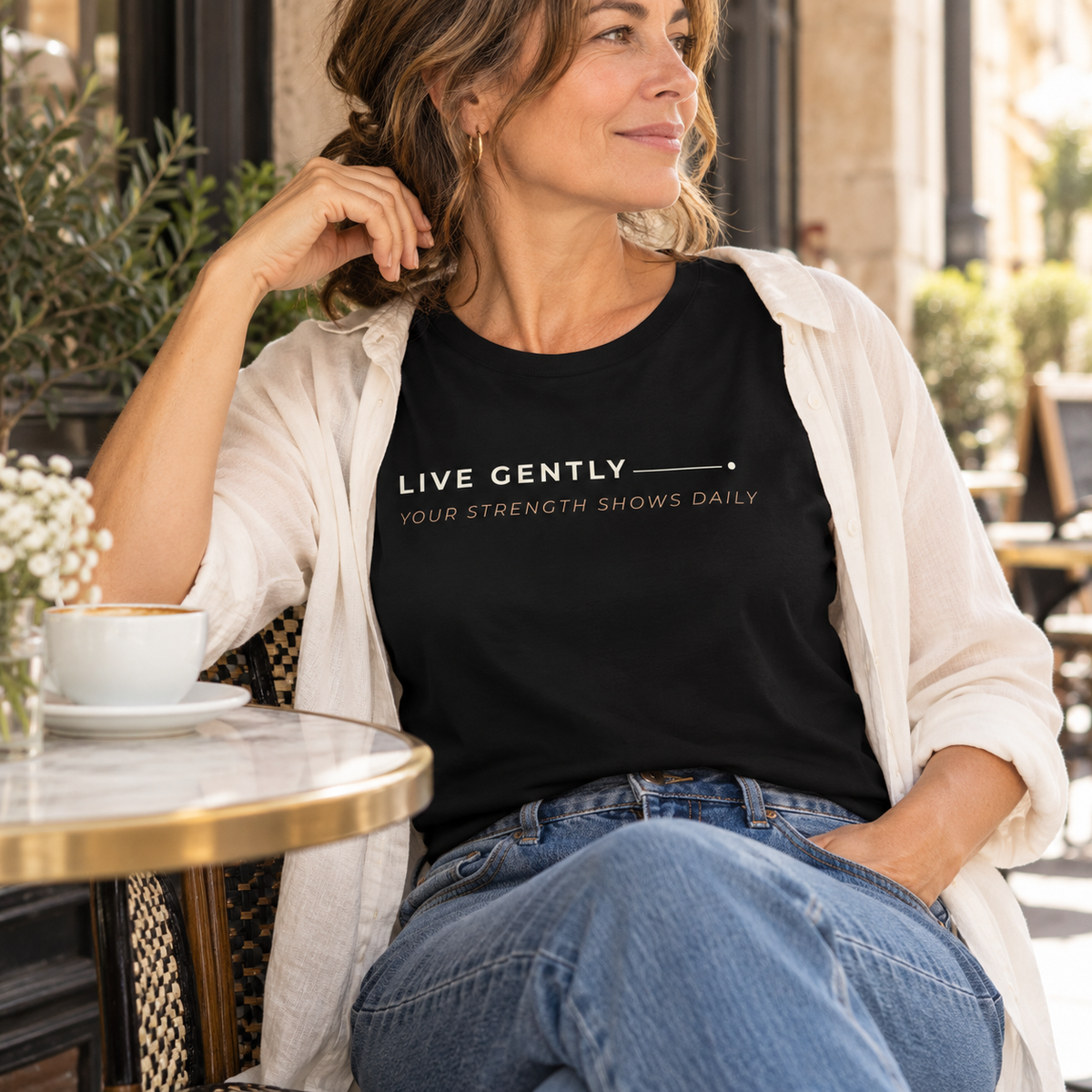 Mulher com T-shirt preta «Live gently, your strength shows daily» e camisa clara aberta, sentada numa esplanada.