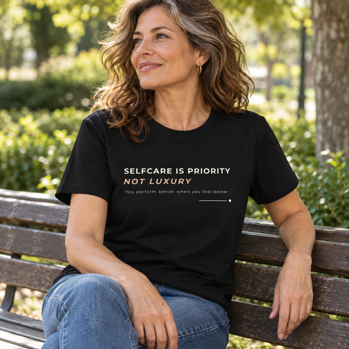 Mulher com T-shirt preta «Selfcare is priority, not luxury» e jeans, sentada num banco de jardim.