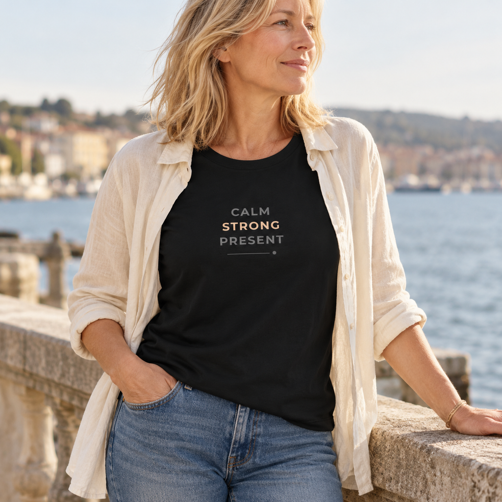 Mulher com T-shirt preta «Calm. Strong. Present.» e camisa clara aberta, junto a um miradouro sobre a água.