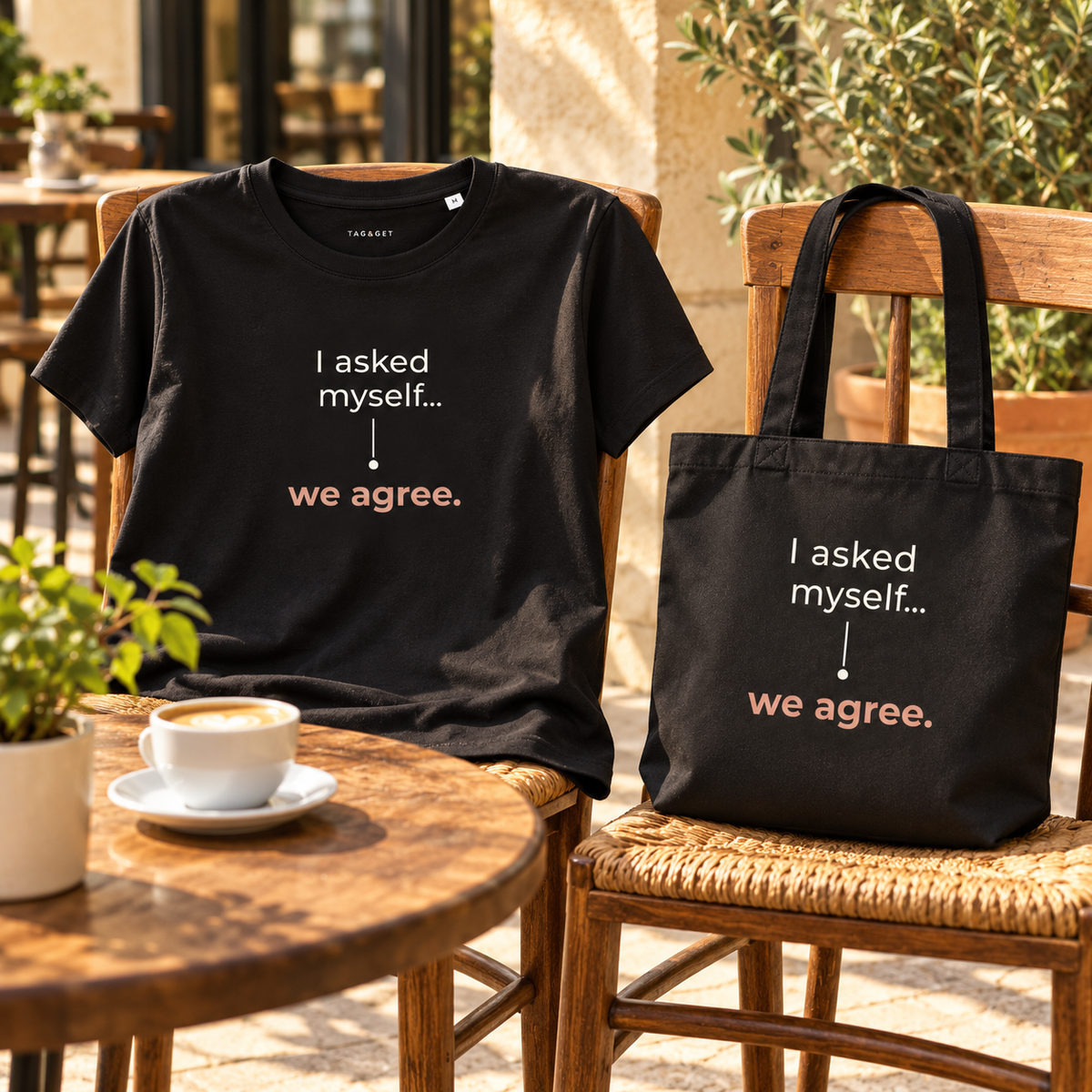 T-shirt e tote bag pretas com a frase “I asked myself... we agree.” em cadeiras de madeira numa esplanada, junto a uma chávena de café.