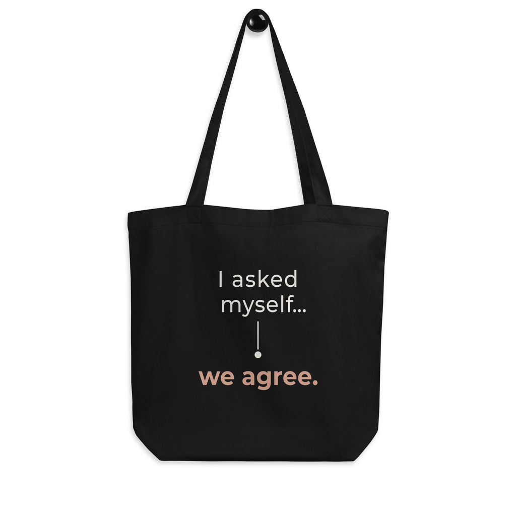 A tote bag preta Tag&Get com a frase “I asked myself... we agree.” em tons claros e pêssego, pendurada pelas alças num gancho sobre fundo branco.