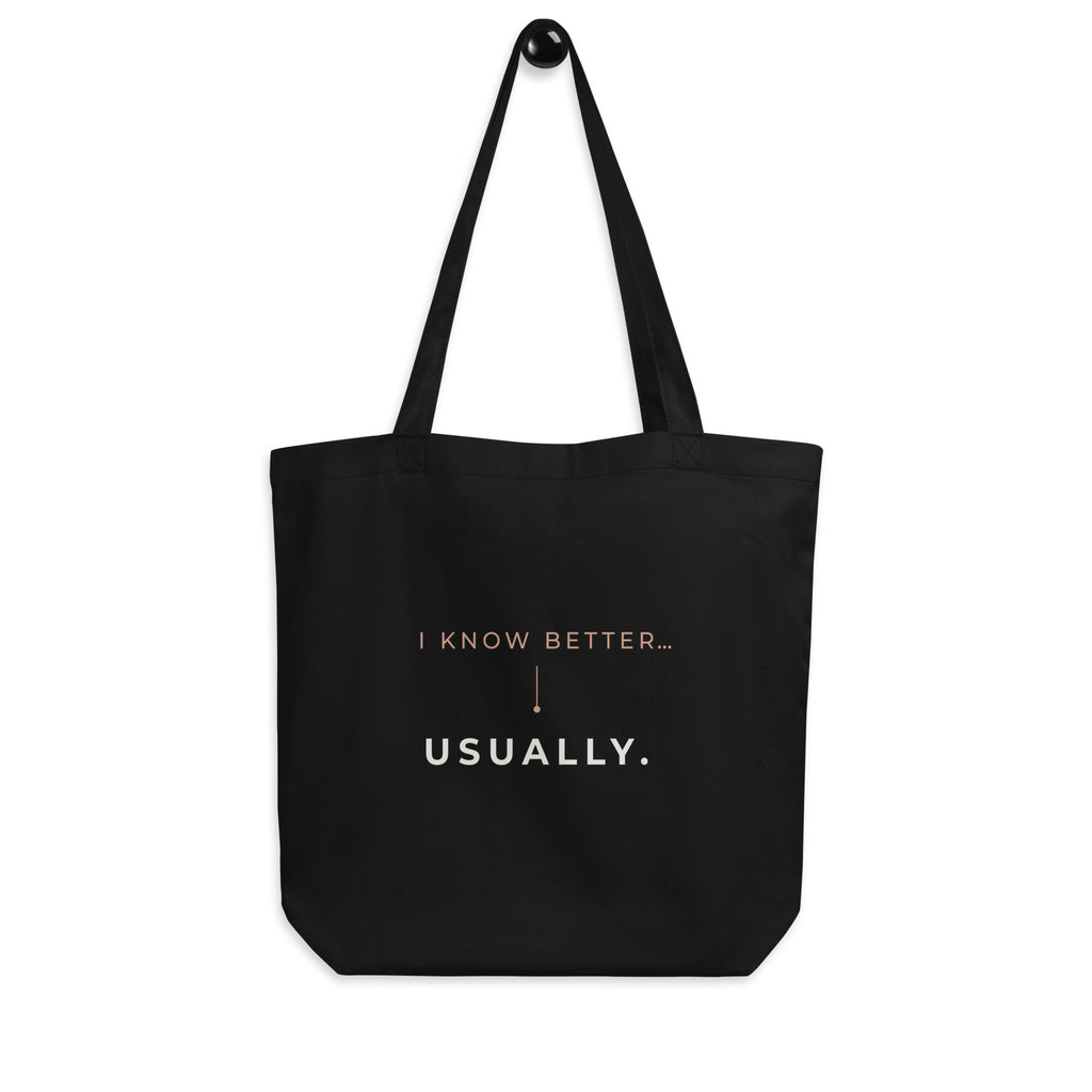 A tote bag preta Tag&Get com a frase “I know better... usually.” em tons claros e pêssego, pendurada pelas alças num gancho sobre fundo branco.
