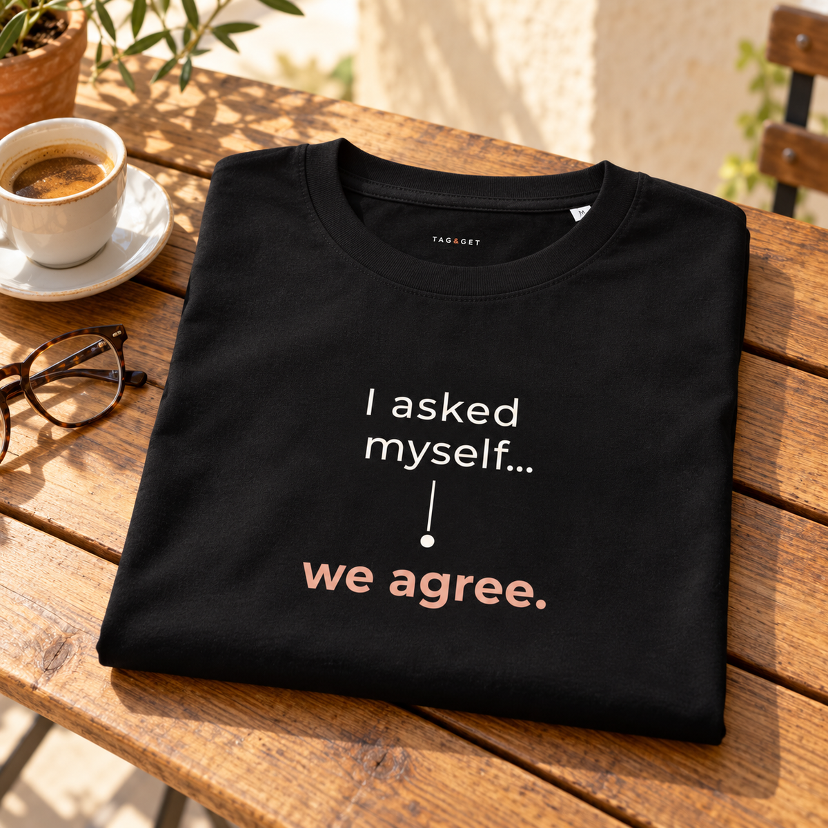 T-shirt preta “I asked myself... we agree.” dobrada sobre uma mesa de madeira, junto a café e óculos numa esplanada.
