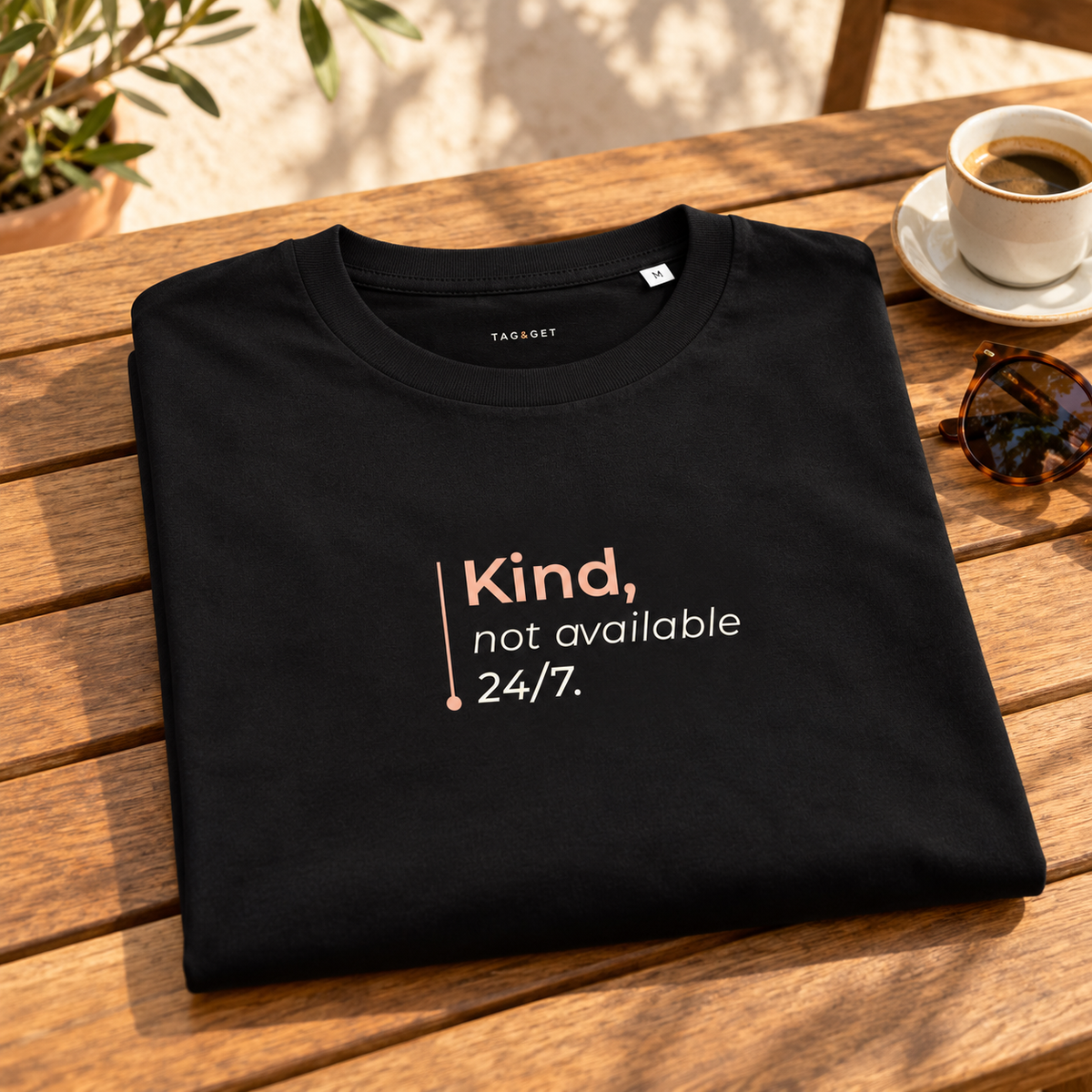 T-shirt preta “Kind, not available 24/7.” dobrada sobre uma mesa de madeira, junto a café e óculos numa esplanada.