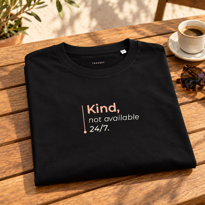 T-shirt preta “Kind, not available 24/7.” dobrada sobre uma mesa de madeira, junto a café e óculos numa esplanada.