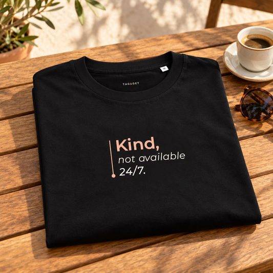 T-shirt preta “Kind, not available 24/7.” dobrada sobre uma mesa de madeira, junto a café e óculos numa esplanada.