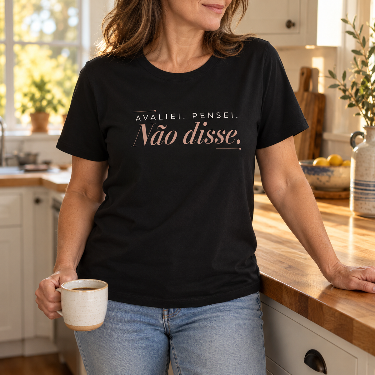 Imagem gerada com estética UGC: mulher com T-shirt preta Avaliei. Pensei. Não disse. com jeans, a segurar uma chávena na cozinha.