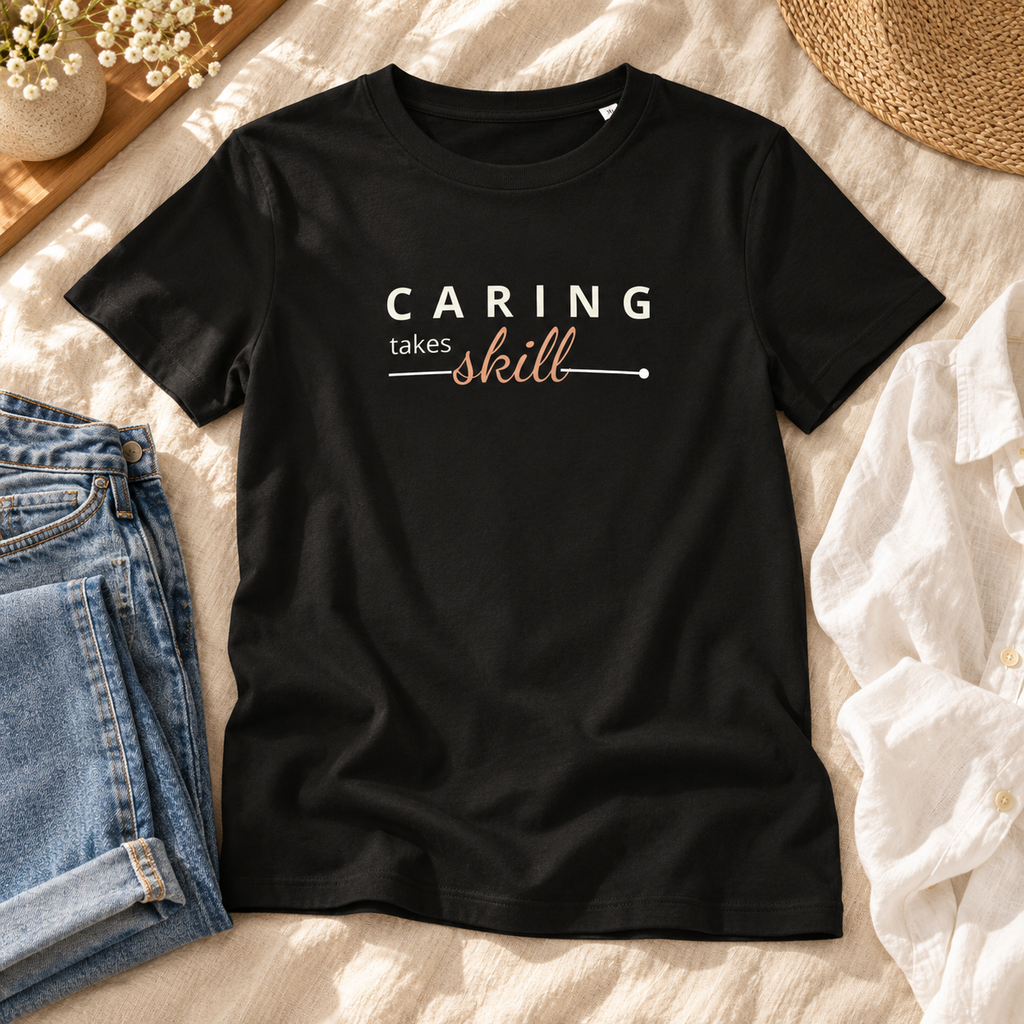 T-shirt preta Caring Takes Skill sobre uma cama de linho claro, ao lado de jeans e camisa branca, com estampado visível.