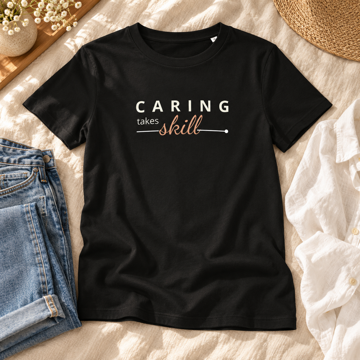 T-shirt preta Caring Takes Skill sobre uma cama de linho claro, ao lado de jeans e camisa branca, com estampado visível.