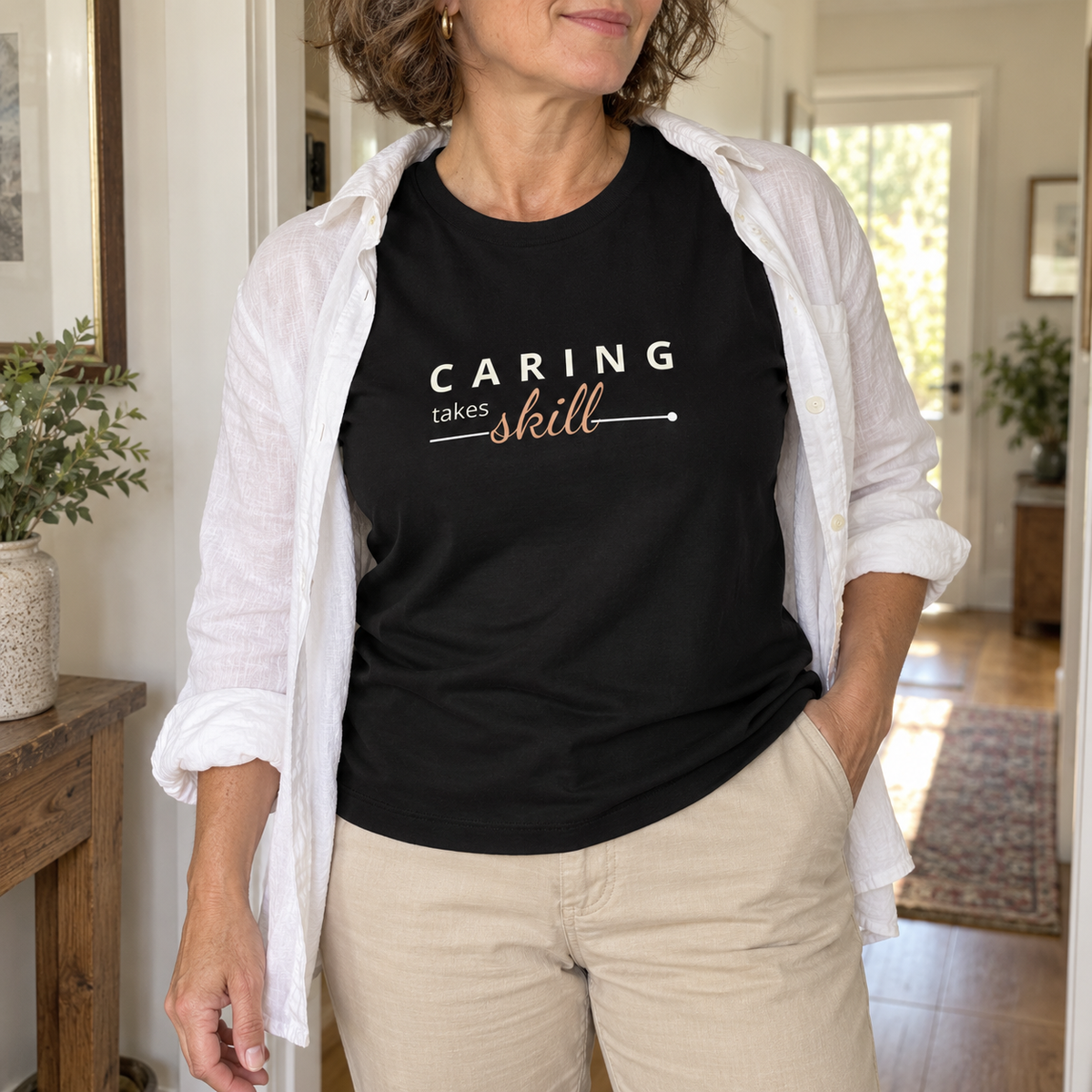 Imagem gerada com estética UGC: mulher com T-shirt preta Caring Takes Skill com calças bege e camisa branca aberta em casa.