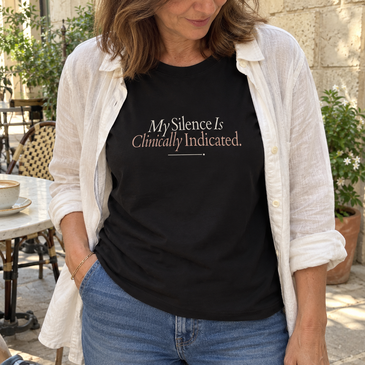 Imagem gerada com estética UGC: mulher com T-shirt preta My Silence Is Clinically Indicated com jeans e camisa branca aberta numa esplanada.