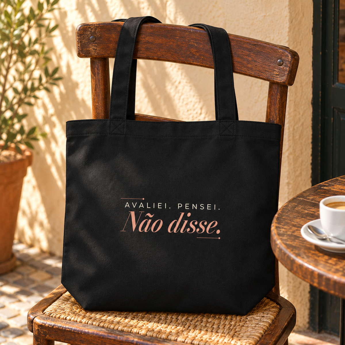 Tote bag preta com a frase “Avaliei. Pensei. Não disse.” numa cadeira de madeira numa esplanada, junto a uma chávena de café.