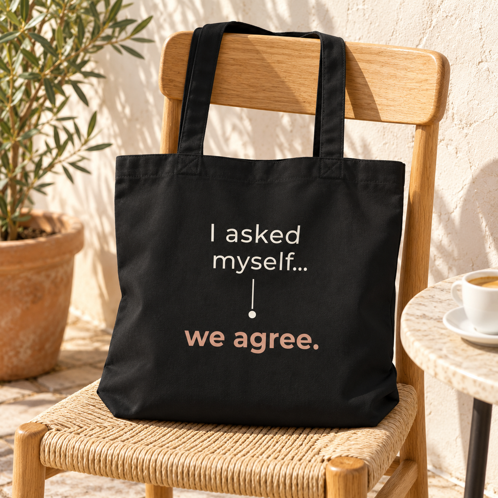 Tote bag preta com a frase “I asked myself... we agree.” numa cadeira de madeira numa esplanada, junto a uma chávena de café.