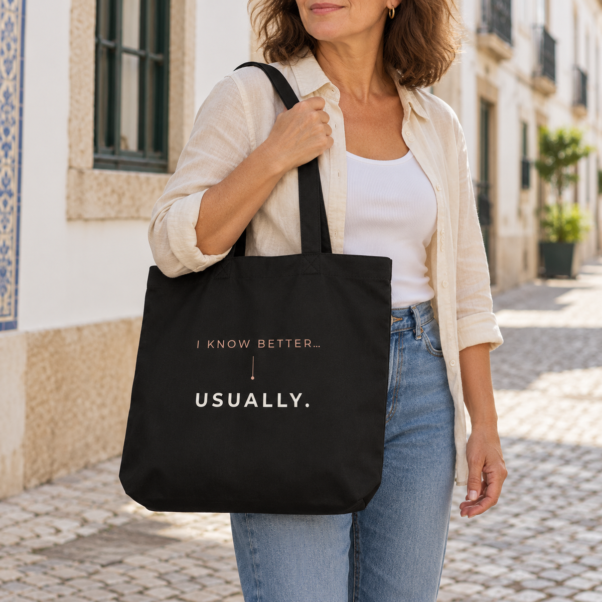 Mulher com camisa clara e jeans a usar ao ombro a tote bag preta com a frase “I know better... usually.” numa rua.