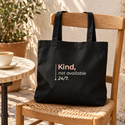 Tote bag preta com a frase “Kind, not available 24/7.” numa cadeira de madeira numa esplanada, junto a uma chávena de café.