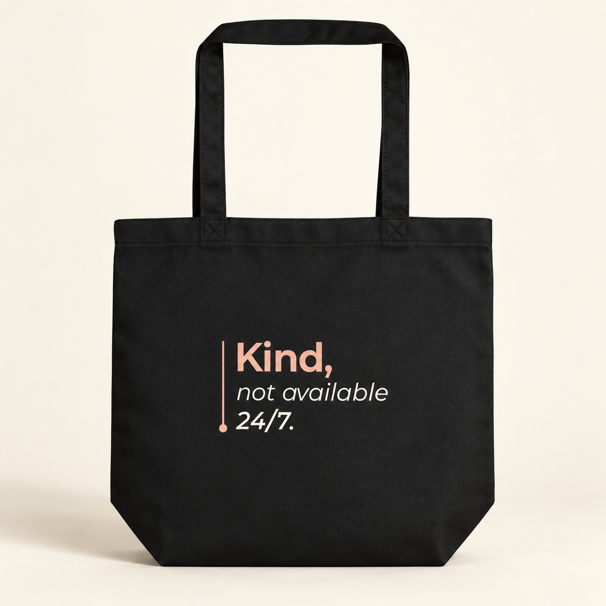 Tote bag preta com a frase “Kind, not available 24/7.”, vista de frente sobre fundo creme.