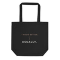 Vista frontal da tote bag preta Tag&Get com a frase “I know better... usually.” em tons claros e pêssego, em algodão orgânico e com alças compridas.