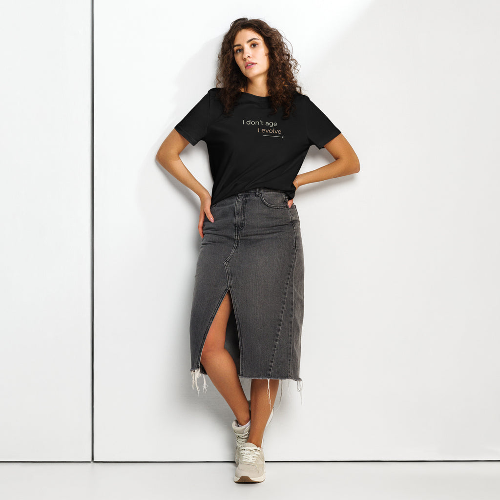 Mulher com T-shirt preta «I don't age, I evolve», saia de ganga escura e ténis, junto a uma parede.