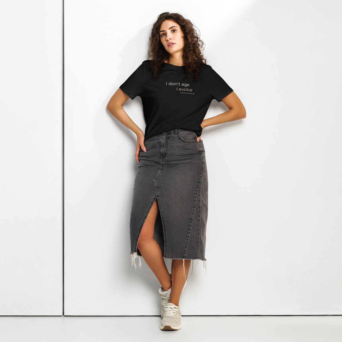 Mulher com T-shirt preta «I don't age, I evolve», saia de ganga escura e ténis, junto a uma parede.